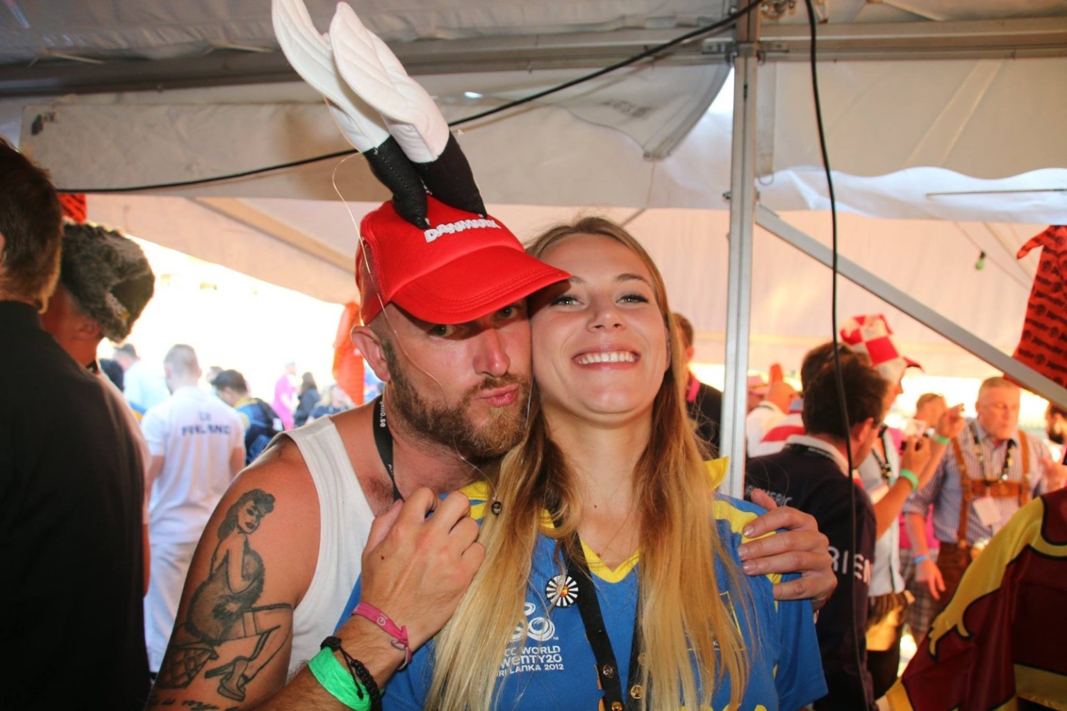 A man in a red novelty cap with tall white ears and a woman in a blue jersey posing cheek to cheek under a tent