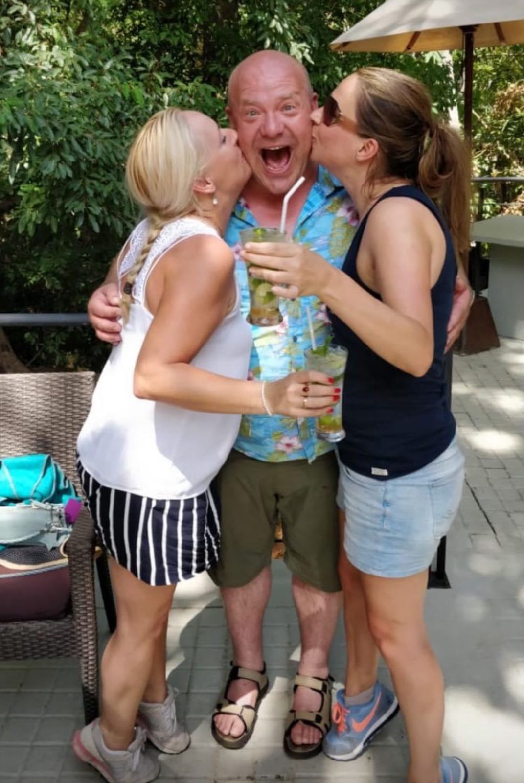 Two friends kissing a laughing man on the cheek, drinks in hand